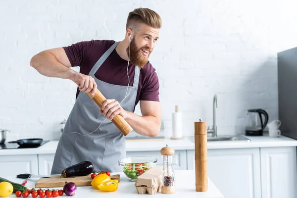 Guapo hombre barbudo sonriente en auriculares cocinar ensalada de verduras y mirando hacia otro lado en la cocina - foto de stock