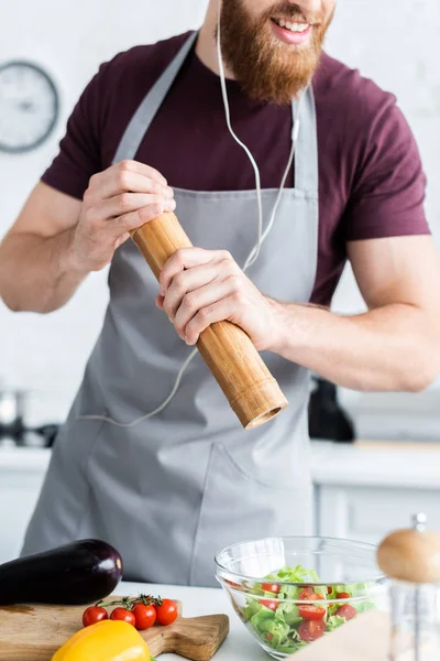 Cropped shot of smiling bearded man in earphones spicing vegetable salad — Stock Photo
