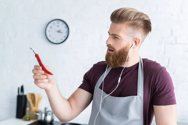 Bonito barbudo jovem em avental e fones de ouvido segurando pimenta na cozinha — Fotografia de Stock