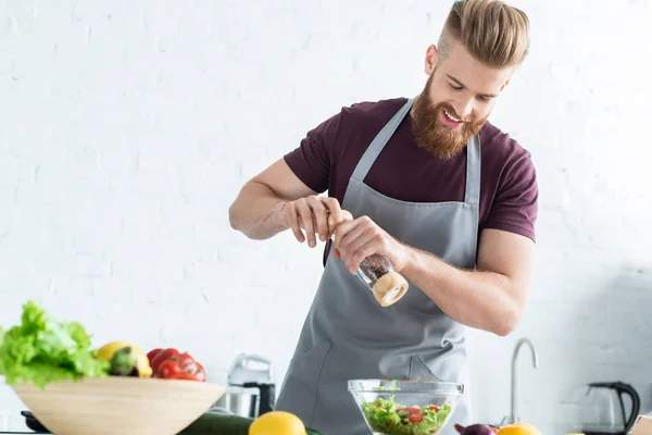 Handsome smiling young man in apron spicing vegetable salad — Stock Photo
