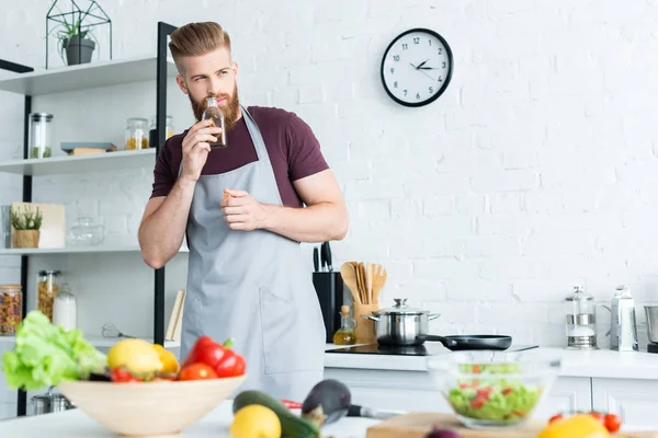 Молодой красивый бородатый мужчина в фартуке смотрит в сторону во время приготовления пищи на кухне — стоковое фото