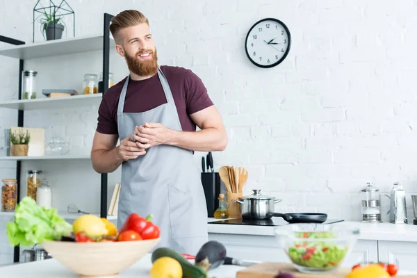 Красивий усміхнений бородатий чоловік в фартусі, дивлячись далеко під час приготування їжі на кухні — стокове фото