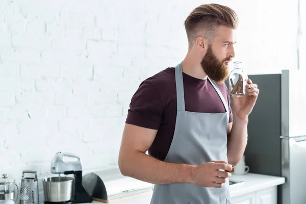 Красивый бородатый молодой человек в фартуке держит контейнер с корицей палочки на кухне — стоковое фото