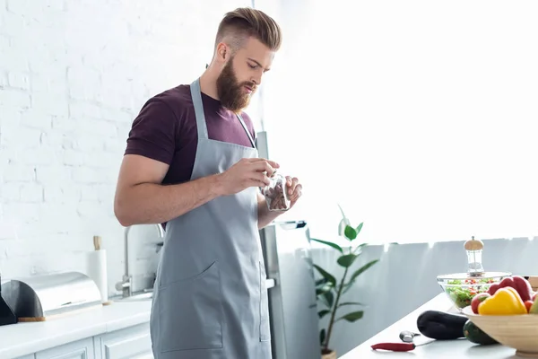 Красивый бородатый мужчина в фартуке держит контейнер с корицей палочки во время приготовления пищи на кухне — стоковое фото