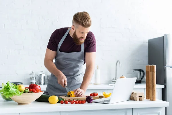 Hombre barbudo guapo en delantal usando portátil y cocinar ensalada de verduras - foto de stock