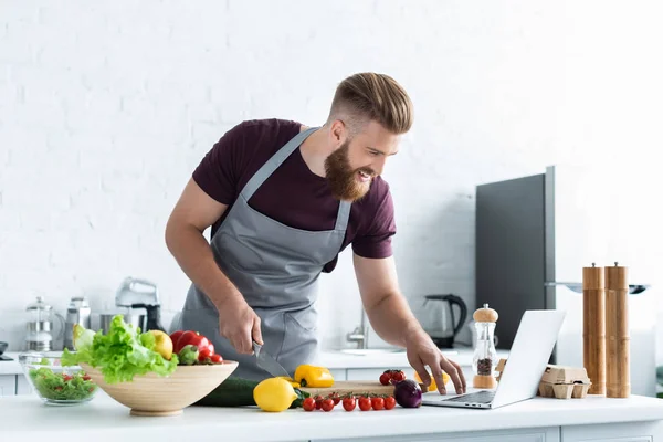 Homem barbudo sorridente em avental usando laptop e cozinhar salada de legumes — Fotografia de Stock