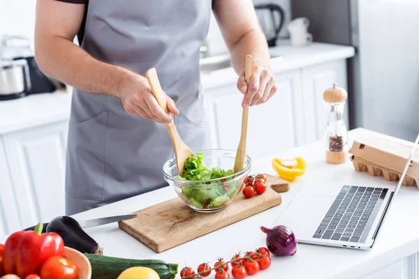 Plan recadré de l'homme dans tablier cuisine salade de légumes et à l'aide d'un ordinateur portable — Photo de stock