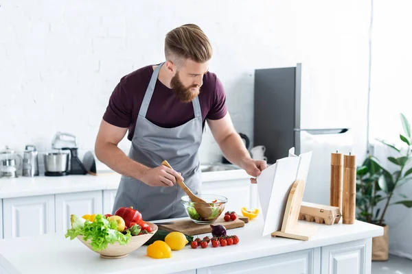 Homem barbudo bonito no avental cozinhar salada de legumes e livro de receitas leitura — Fotografia de Stock