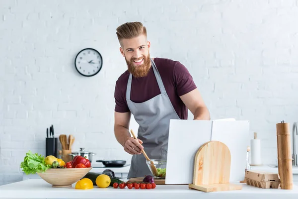 Bel homme barbu dans tablier cuisine salade de légumes et souriant à la caméra — Photo de stock