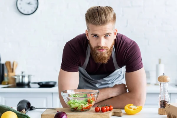 Красивий бородатий чоловік в фартусі готує овочевий салат і дивиться на камеру — стокове фото