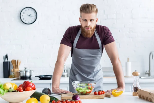 Guapo barbudo hombre en delantal cocinar ensalada de verduras y mirando a la cámara - foto de stock