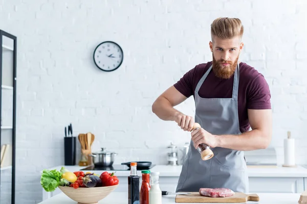 Schöner bärtiger Mann in Schürze kocht leckeres Steak und schaut in die Kamera — Stockfoto