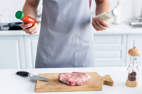 Cropped shot of man in apron holding sauces while cooking beef steak — Stock Photo