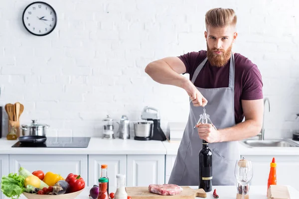 Schöner bärtiger junger Mann in Schürze öffnet eine Flasche Wein und schaut in die Kamera, während er in der Küche kocht — Stockfoto