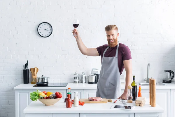 Schöner bärtiger junger Mann in Schürze hält ein Glas Rotwein in der Hand und lächelt in die Kamera, während er in der Küche kocht — Stockfoto