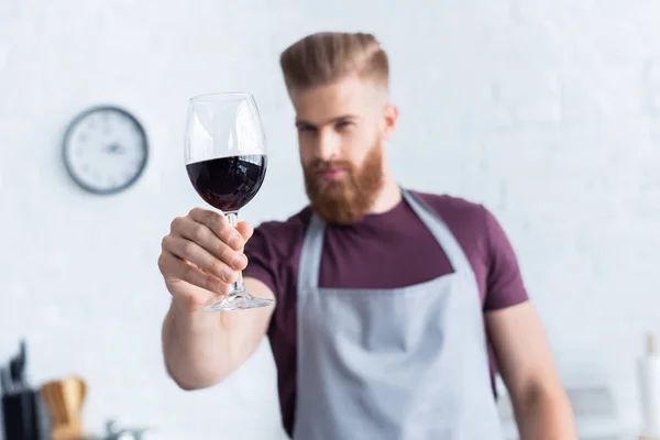 Bel giovane barbuto in grembiule con in mano un bicchiere di vino rosso in cucina — Foto stock