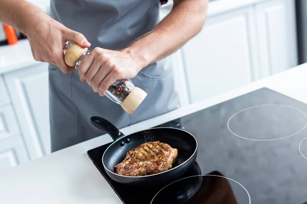 Tiro recortado de hombre sosteniendo molino de pimienta mientras cocina delicioso bistec en sartén - foto de stock