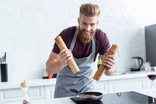 Handsome bearded young man in apron smiling at camera while cooking delicious beef steak in kitchen — Stock Photo