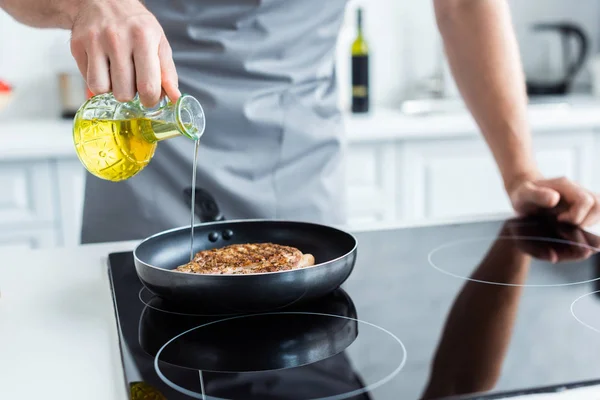 Colpo ritagliato di uomo in grembiule versando olio durante la cottura bistecca sulla padella — Foto stock