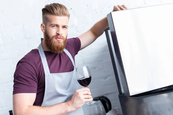 Hombre barbudo guapo en delantal sosteniendo copa de vino tinto y mirando a la cámara - foto de stock