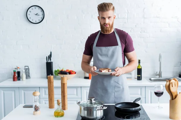 Handsome bearded young man in apron holding delicious beef steak on plate and looking at camera — Stock Photo