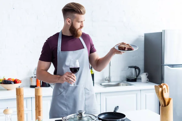 Handsome young man in apron holding glass of red wine and delicious steak on plate — Stock Photo