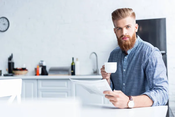 Schöner bärtiger junger Mann hält eine Tasse Kaffee in der Hand und schaut in die Kamera, während er zu Hause Zeitung liest — Stockfoto