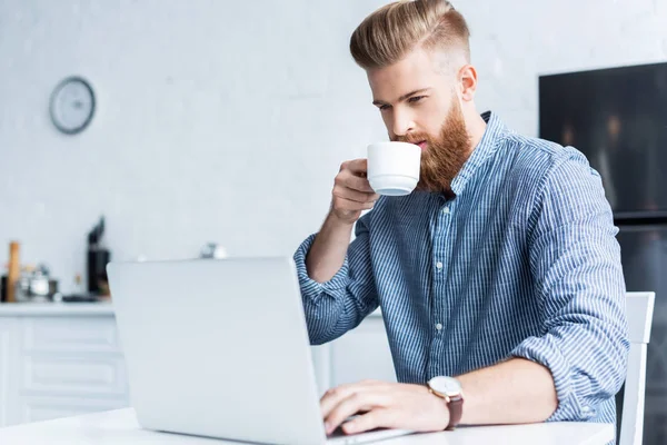 Bonito barbudo jovem bebendo café e usando laptop em casa — Fotografia de Stock