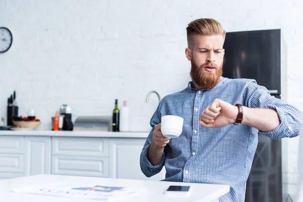 Beau jeune homme barbu tenant tasse de café et de vérifier montre-bracelet tout en étant assis à la table — Photo de stock