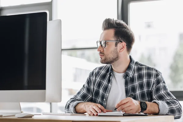 Handsome young programmer in eyeglasses taking notes and working with desktop computer — Stock Photo