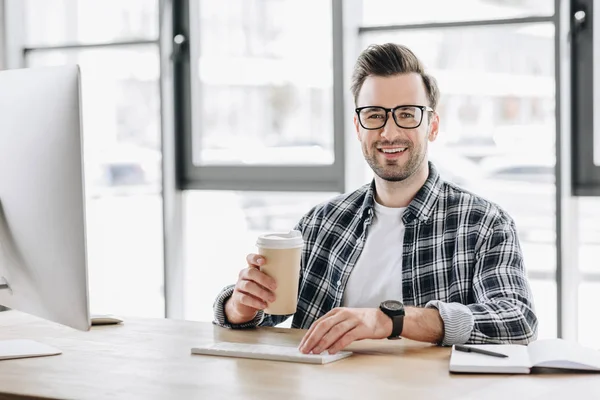 Guapo joven programador sosteniendo taza de papel y sonriendo a la cámara mientras está sentado en el lugar de trabajo - foto de stock