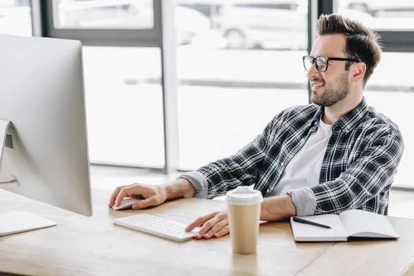 Jovem sorridente em óculos usando computador desktop no local de trabalho — Fotografia de Stock