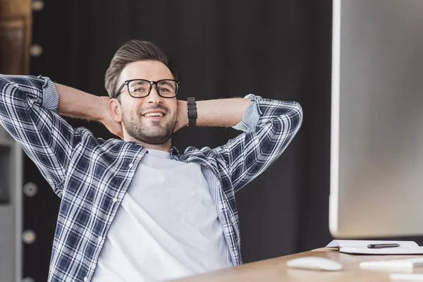 Guapo joven programador en anteojos sonriendo a la cámara mientras está sentado con las manos detrás de la cabeza en el lugar de trabajo - foto de stock