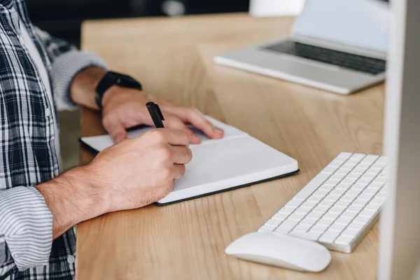 Recortado tiro del hombre tomando notas en el bloc de notas mientras se utiliza la computadora de escritorio en el lugar de trabajo - foto de stock