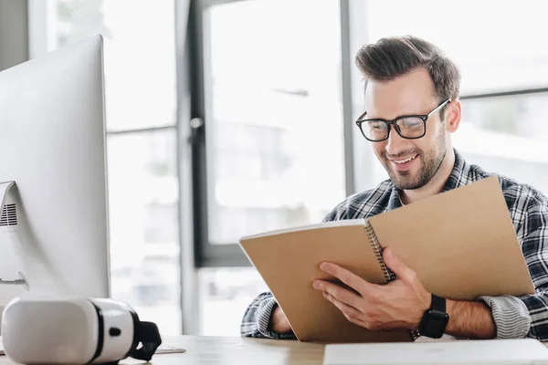 Jeune homme souriant dans des lunettes écrit dans un cahier sur le lieu de travail — Photo de stock