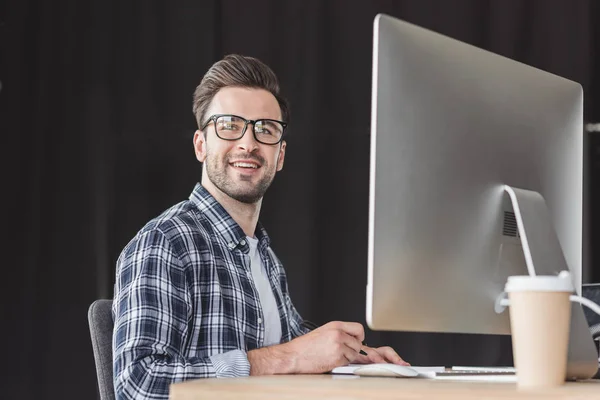 Handsome smiling young programmer in eyeglasses working with desktop computer — Stock Photo