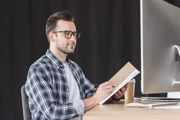 Schöner junger Programmierer mit Brille, der Notizen im Notizbuch macht und Desktop-Computer benutzt — Stockfoto