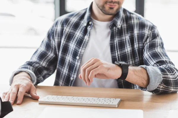 Corte tiro de jovem verificando smartwatch enquanto sentado no local de trabalho — Fotografia de Stock