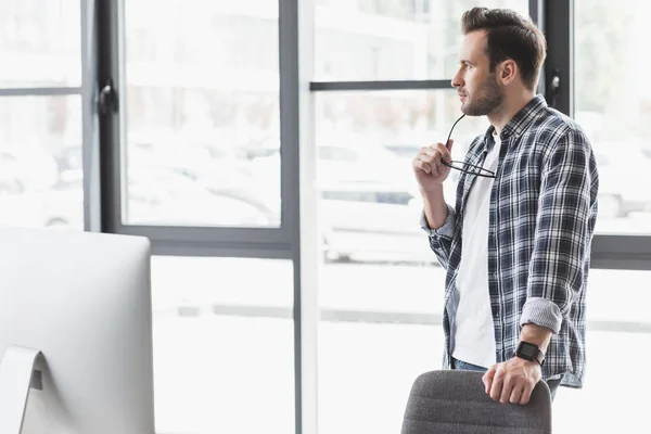 Pensive young man holding eyeglasses and looking away while standing at workplace — Stock Photo