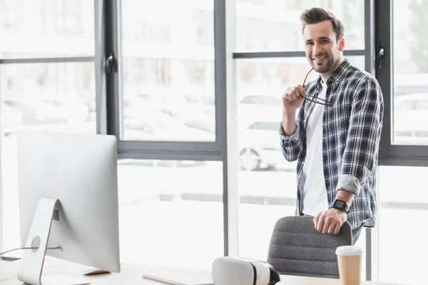 Bello giovane programmatore che tiene gli occhiali e sorride alla fotocamera mentre si trova sul posto di lavoro — Foto stock