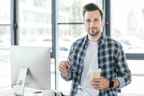 Young man looking at camera while holding eyeglasses and paper cup at workplace — Stock Photo