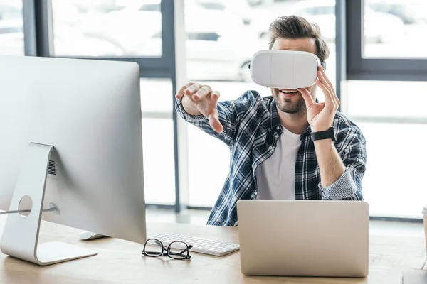 Улыбающийся молодой человек в наушниках виртуальной реальности с помощью ноутбука и настольного компьютера на рабочем месте — стоковое фото