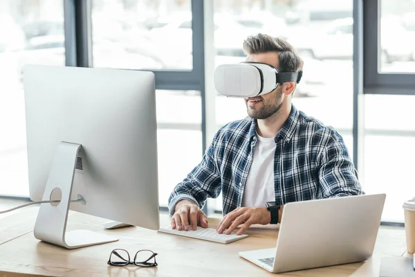 Hombre joven con auriculares de realidad virtual usando computadora de escritorio y computadora portátil en el lugar de trabajo - foto de stock