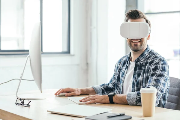 Улыбающийся молодой человек в наушниках виртуальной реальности за настольным компьютером на рабочем месте — стоковое фото
