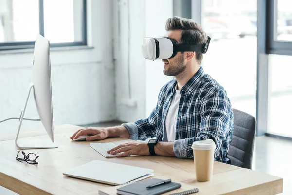 Hombre joven con auriculares de realidad virtual usando computadora de escritorio en el lugar de trabajo - foto de stock