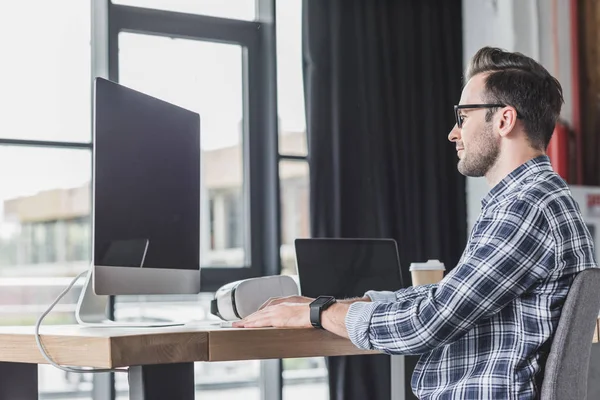 Schöner lächelnder junger Programmierer mit Brille, der mit Desktop-Computer und Laptop arbeitet — Stockfoto