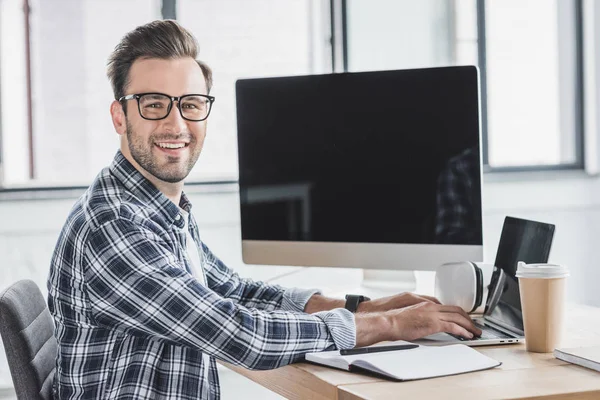 Schöner junger Programmierer mit Brille, der während der Arbeit mit Laptop und Desktop-Computer in die Kamera lächelt — Stockfoto