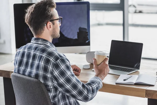 Joven con gafas de vista sosteniendo una taza de papel mientras trabaja con una computadora portátil y de escritorio - foto de stock