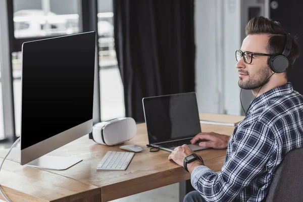 Beau jeune programmeur souriant dans les écouteurs et les lunettes à l'aide d'un ordinateur portable et ordinateur de bureau — Photo de stock