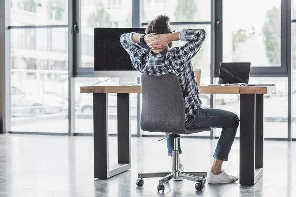 Rückansicht eines jungen Programmierers, der mit den Händen hinter dem Kopf am Arbeitsplatz sitzt — Stockfoto